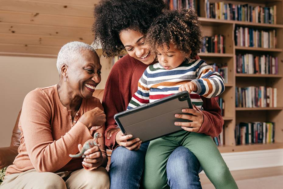 A grandmother smiles and sits with her daughter and granddaughter while looking at a tablet together.