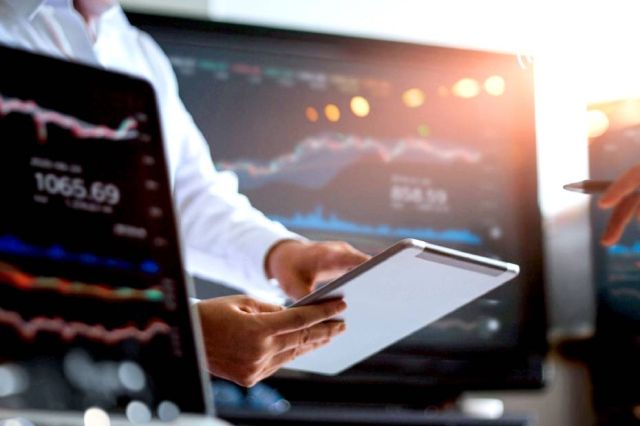 A person holds a tablet among computers displaying financial graphs.