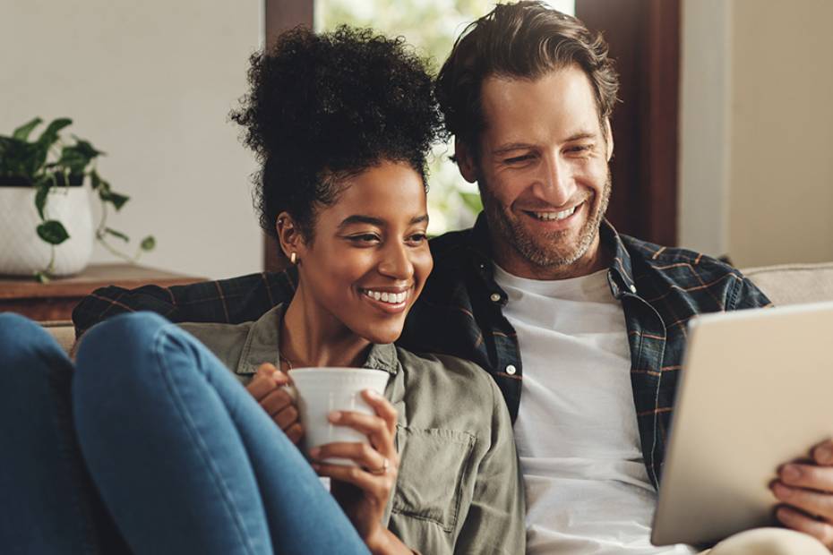 A picture showing a happy couple relaxing on sofa, looking at something on the tablet he is holding.