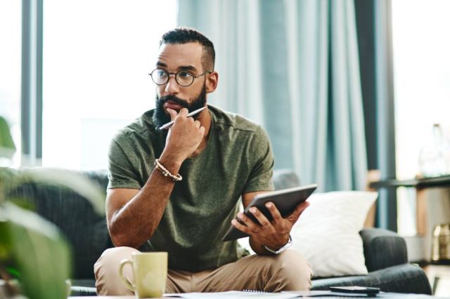 A picture showing a young guy sitting on a sofa in thoughts, with a pen and tablet in his hands.