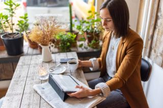 Woman wearing a blazer holding a coffee mug and reading news on a tablet with a newspaper on a wooden desk.