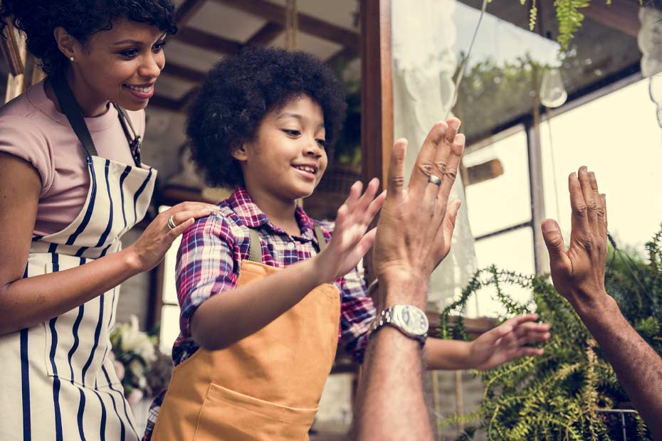 Smiling woman stands behind child wearing apron who is high fiving a man.