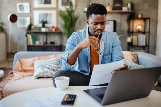 Homme lisant une feuille de papier devant un ordinateur portable allumé.