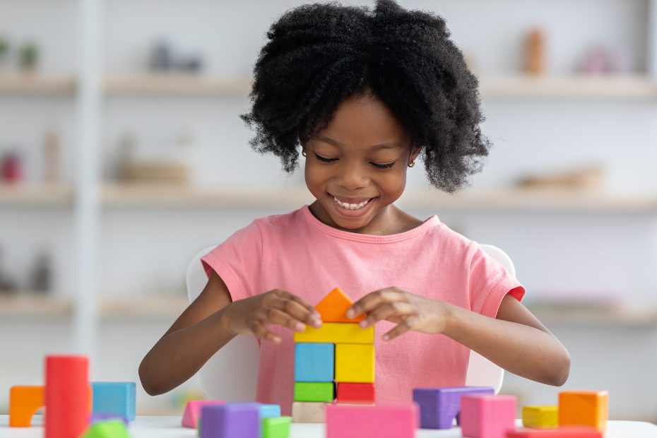 Fillette afro-américaine, souriante, vêtue d'un tee-shirt rose, jouant avec des cubes de bois colorés.