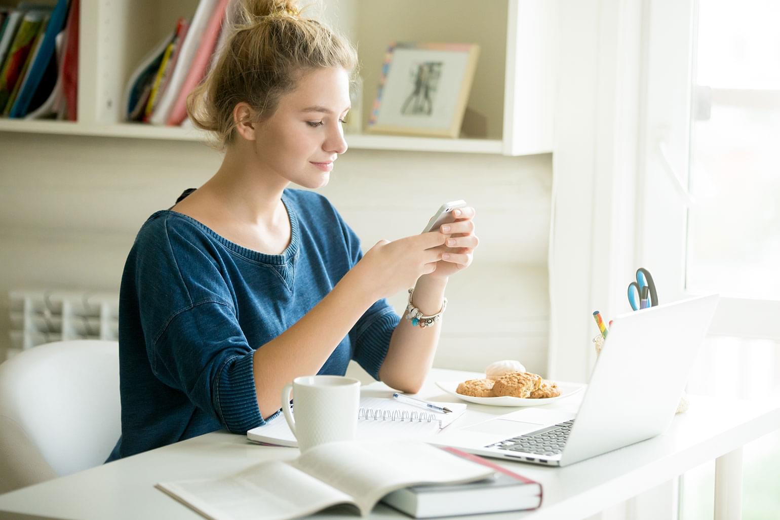 Portrait d’une belle jeune femme décontractée tenant un téléphone intelligent. Assise derrière un bureau moderne, à la maison ou dans une résidence étudiante, et entourée d’un ordinateur portable, de livres, d’un café et de biscuits, elle regarde l’écran et utilise une application ou un service de messagerie texte.
